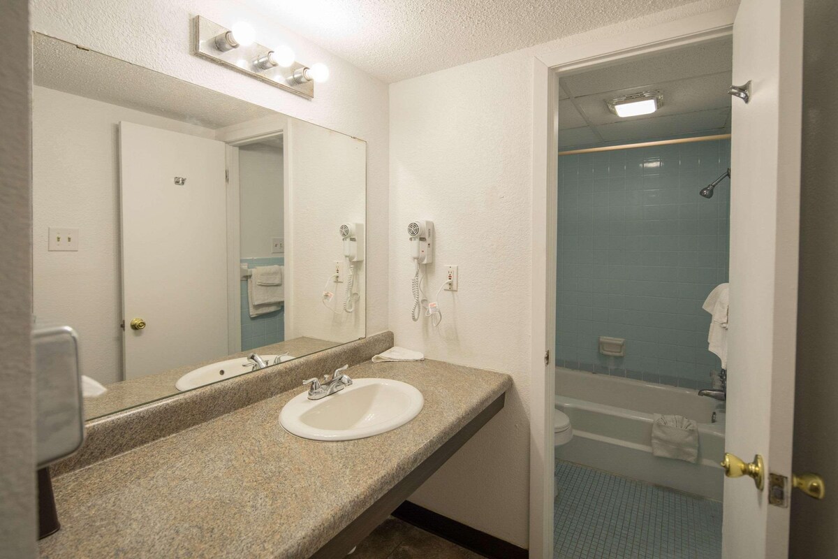 A bathroom with a granite countertop, a white sink, a mirror, a hairdryer, and a shower area.