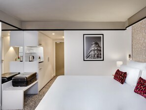 A hotel room with a bed, a desk with a laptop, a coffee machine, and a framed picture on the wall.