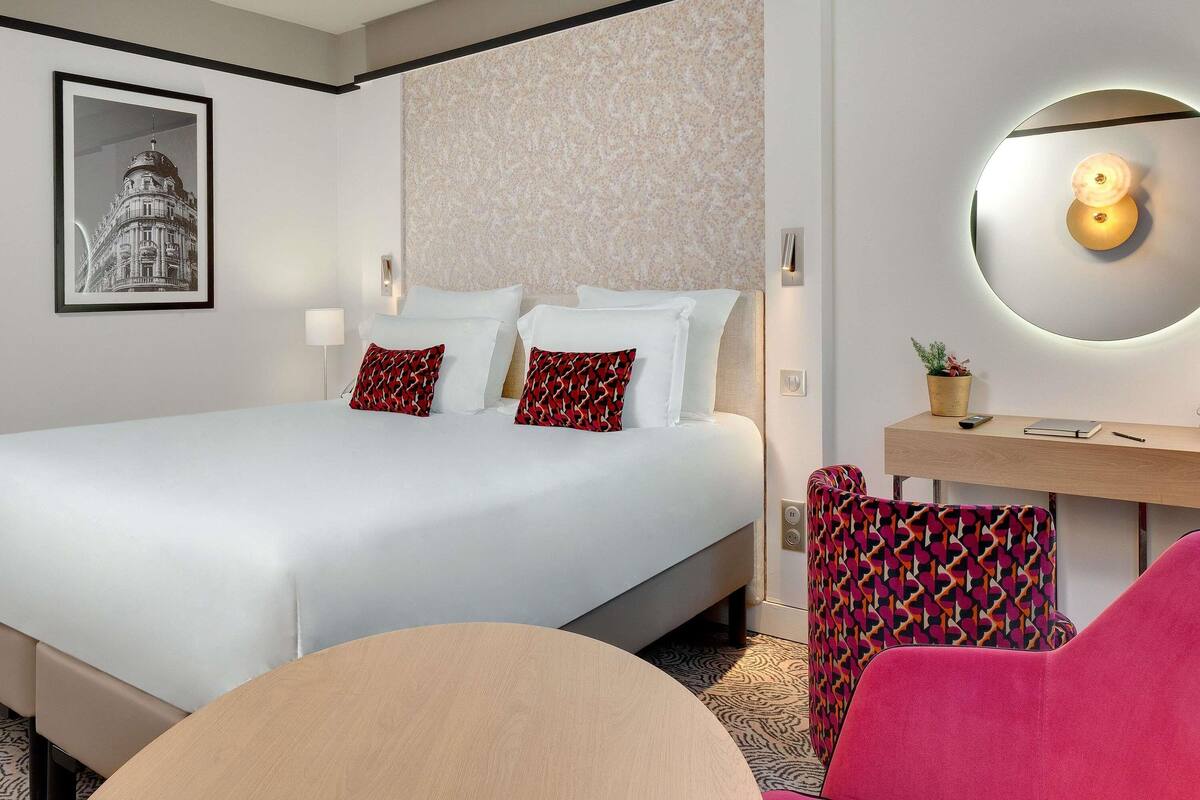 Quarto de hotel com uma cama grande, uma escrivaninha com abajur, uma cadeira e um quadro na parede.