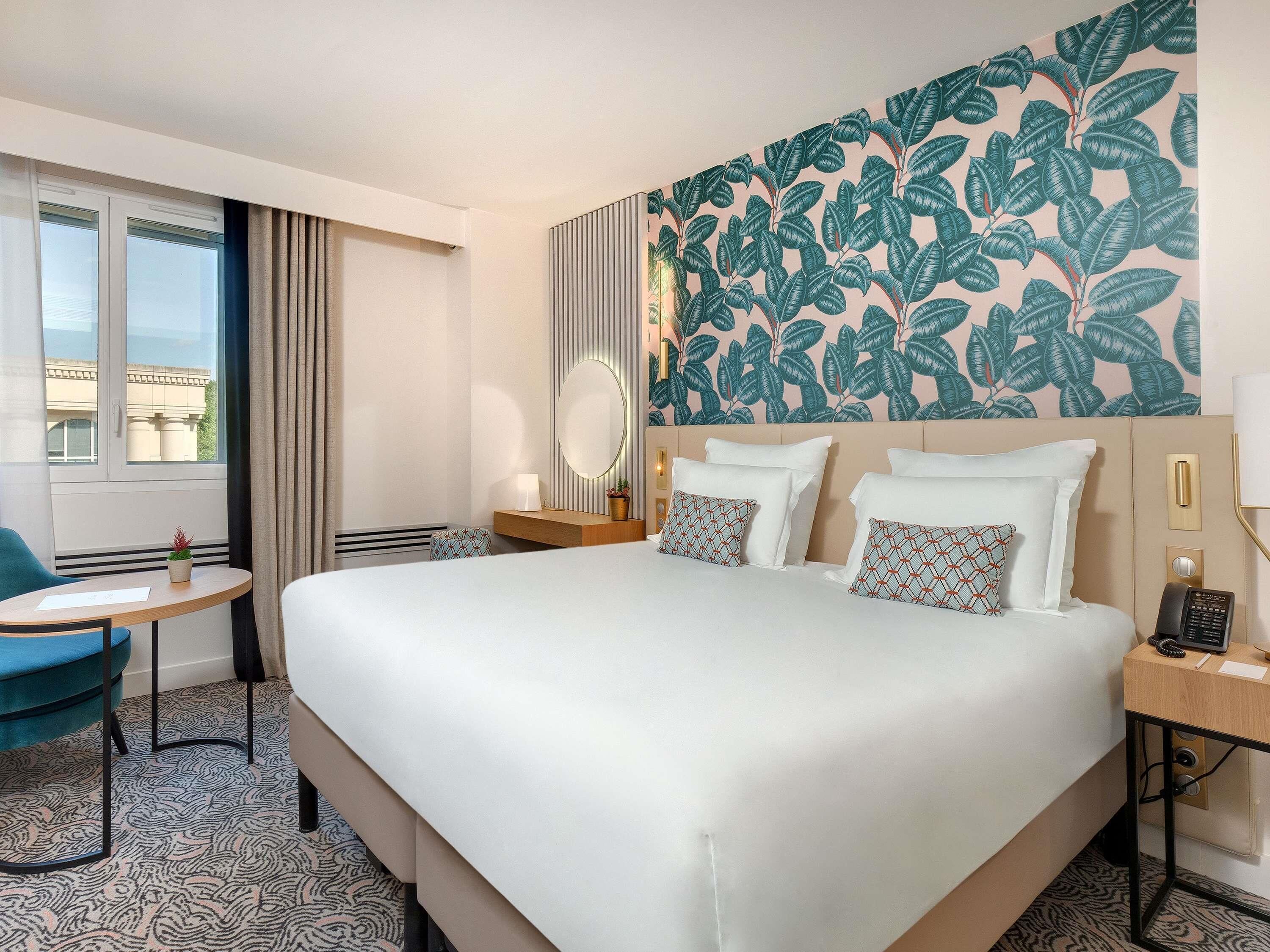 A hotel room with a large bed, a desk, a chair, a window, and a wall with a leaf pattern.