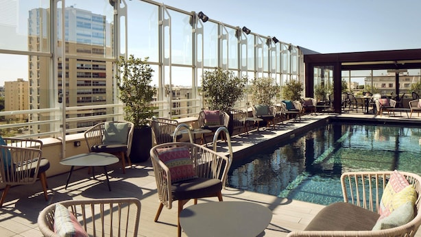 A rooftop terrace with a pool, wicker furniture, and large windows offering city views.