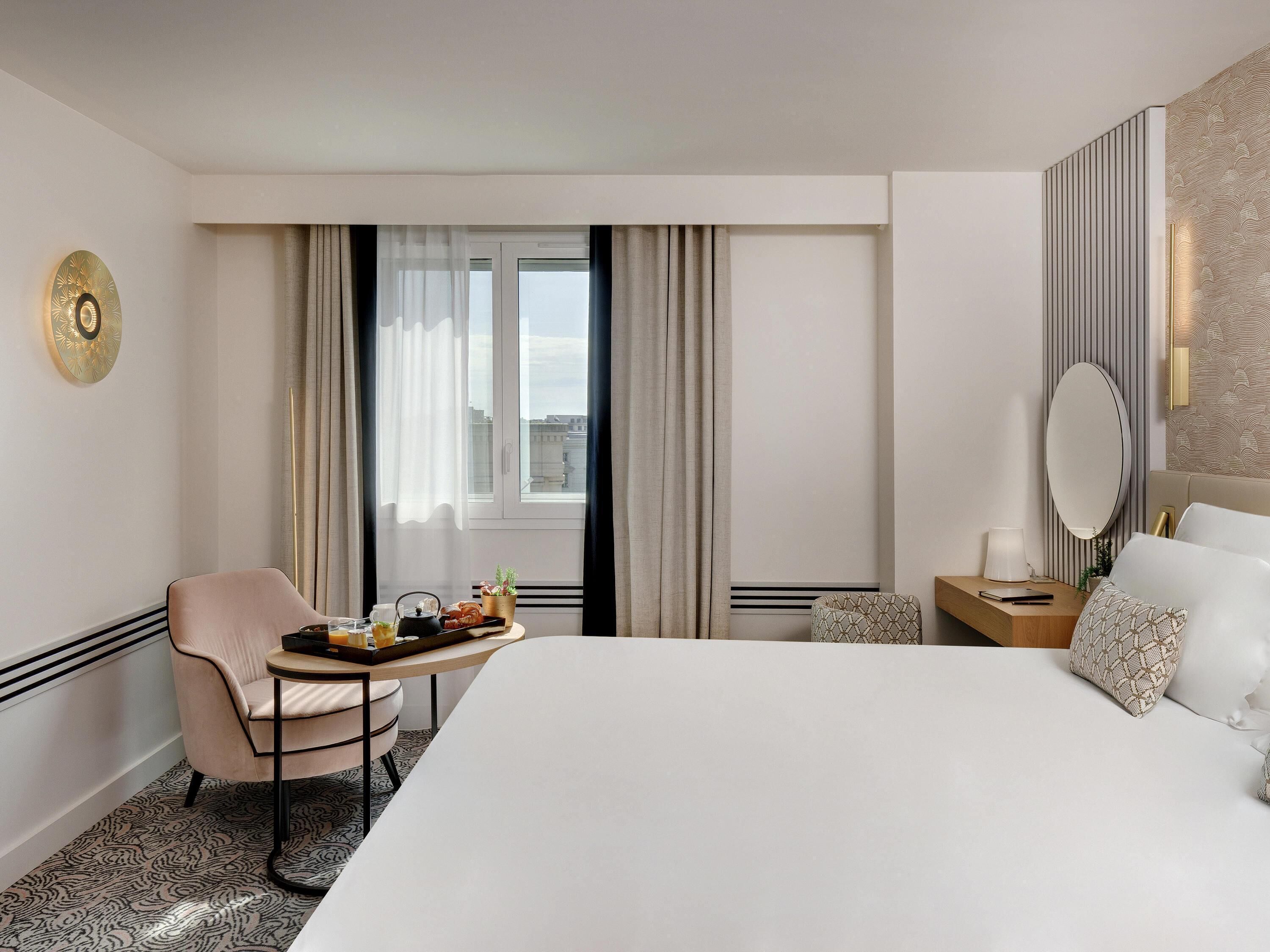 A hotel room with a bed, a chair, a small table with breakfast items, a bedside table with a lamp, and a large window with curtains.