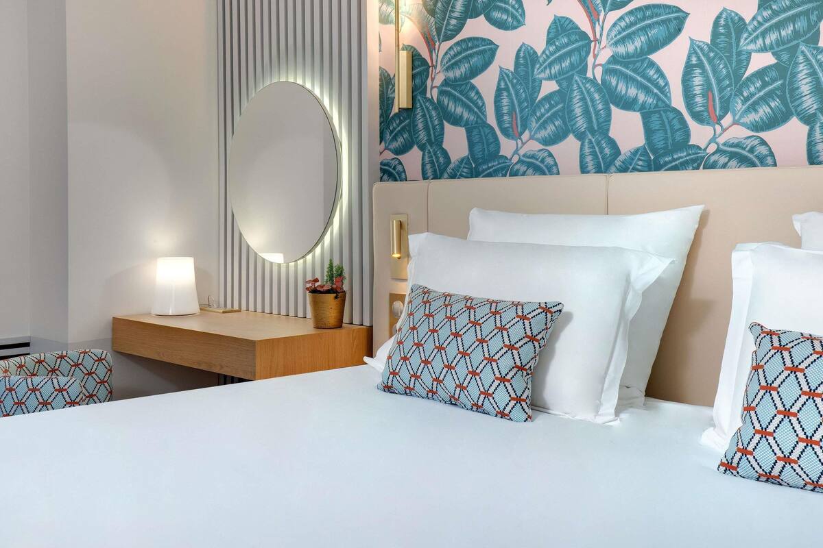 Quarto de hotel com cama, mesa de cabeceira, luminária, espelho e parede com padrão de folhas.