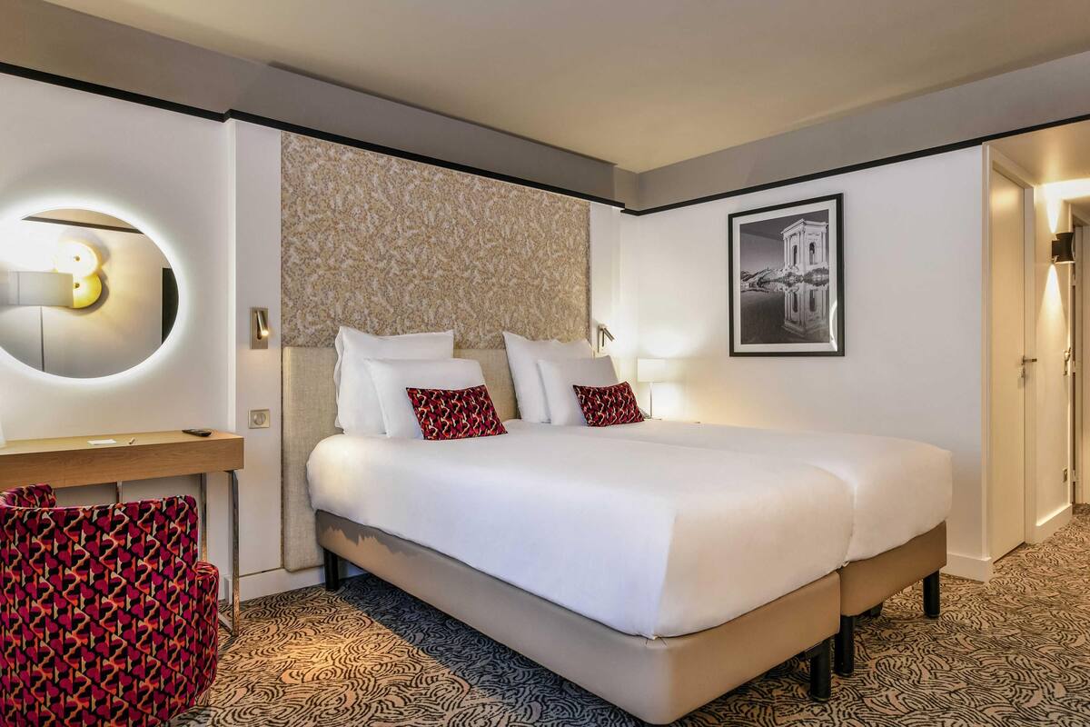 Quarto de hotel com uma cama grande, uma mesinha de cabeceira, um espelho, um tapete estampado e um quadro na parede.