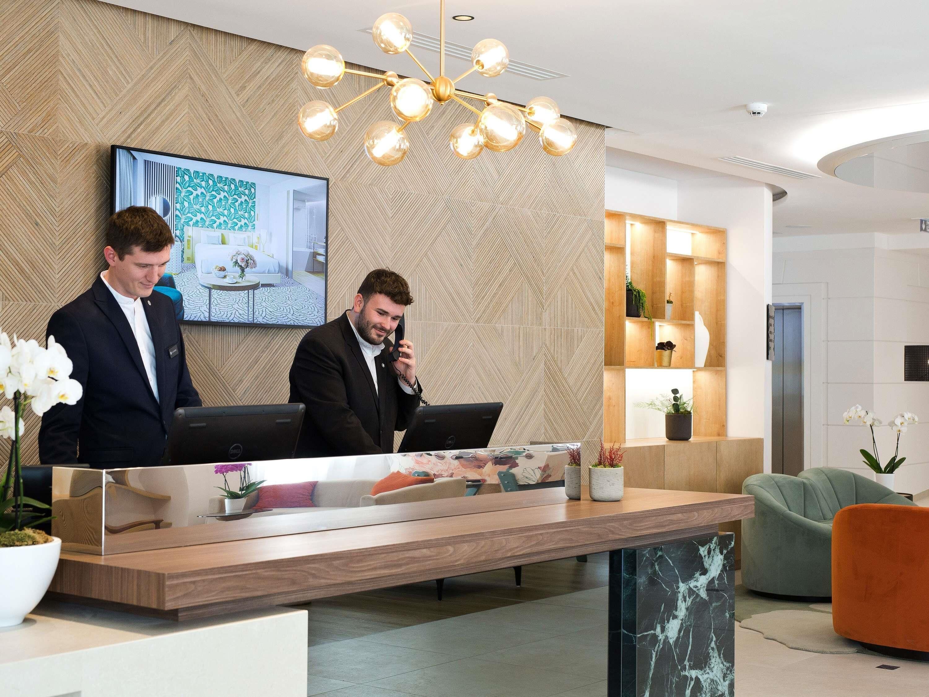 Um balcão de atendimento moderno com dois computadores, uma tela grande exibindo um quarto, e um elegante candelabro.