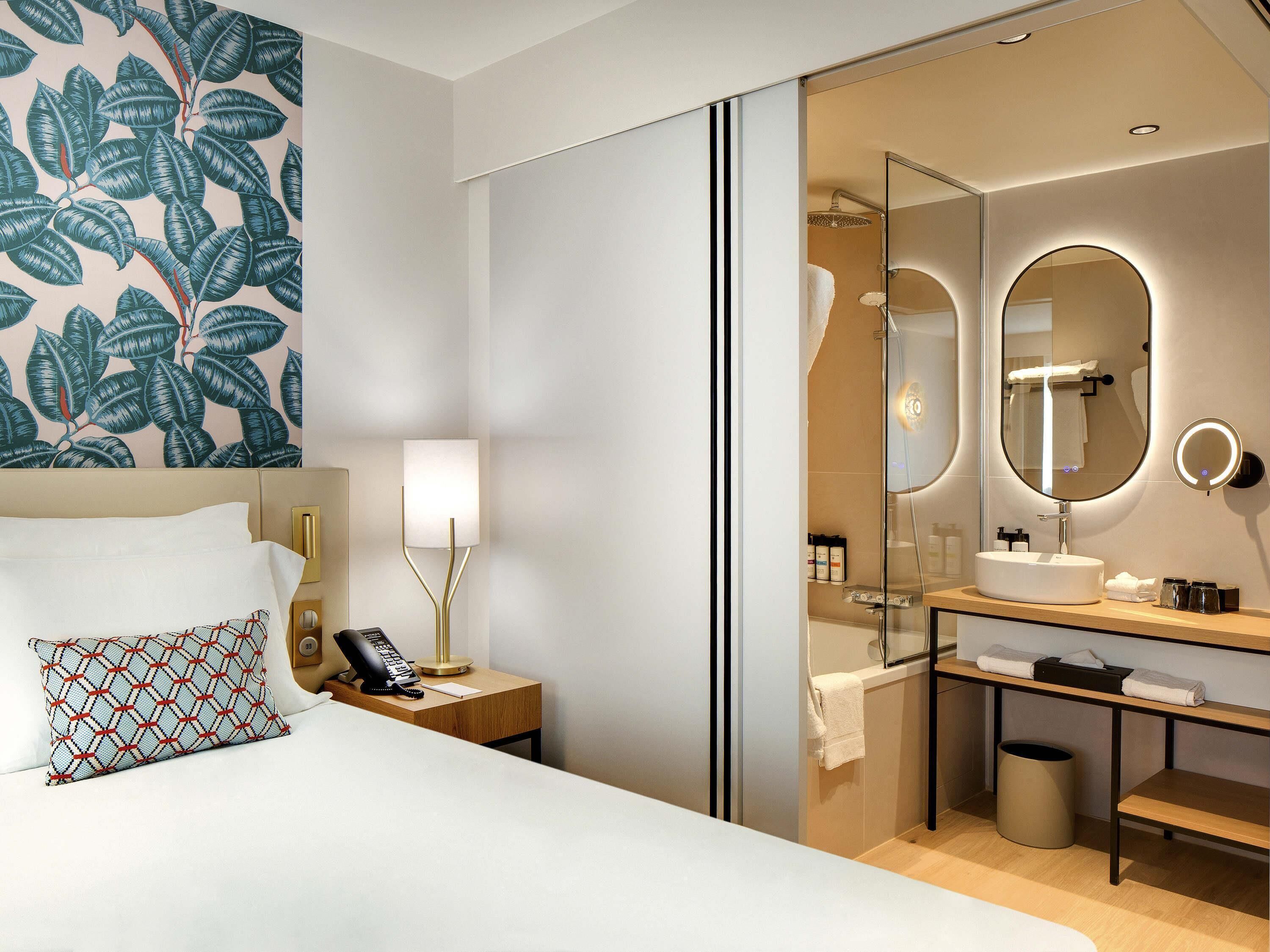 Ein Hotelzimmer mit einem Bett, einem Nachttisch mit Lampe, einem Telefon, einem Spiegel und einem Badezimmer mit Waschbecken, Spiegel und Handtuchhalter.