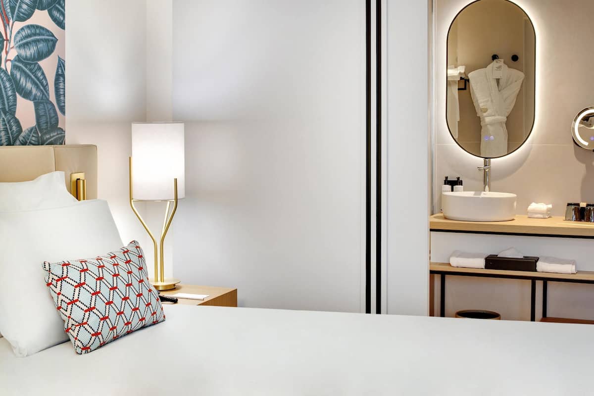 Quarto de hotel com cama, luminária, mesa de cabeceira, espelho, pia e porta-toalhas.