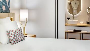 Habitación de hotel con cama, lámpara de noche, mesita de noche, espejo, lavamanos y perchero.