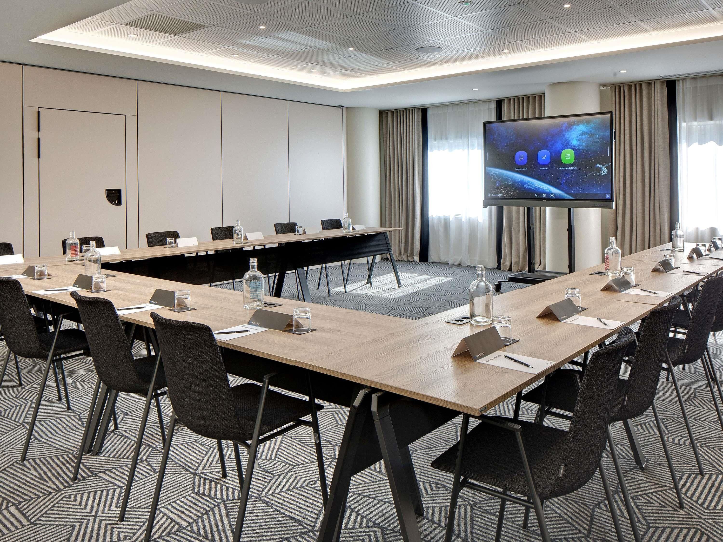 Un salón de conferencias con una larga mesa, sillas, una pantalla de proyector y un amplio ventanal con cortinas.