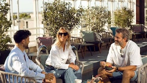 Three people seated at an outdoor café with tables and chairs, surrounded by greenery and a view of buildings.
