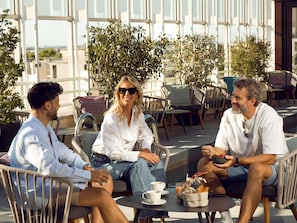 Three people seated at an outdoor café with tables and chairs, surrounded by greenery and a view of buildings.