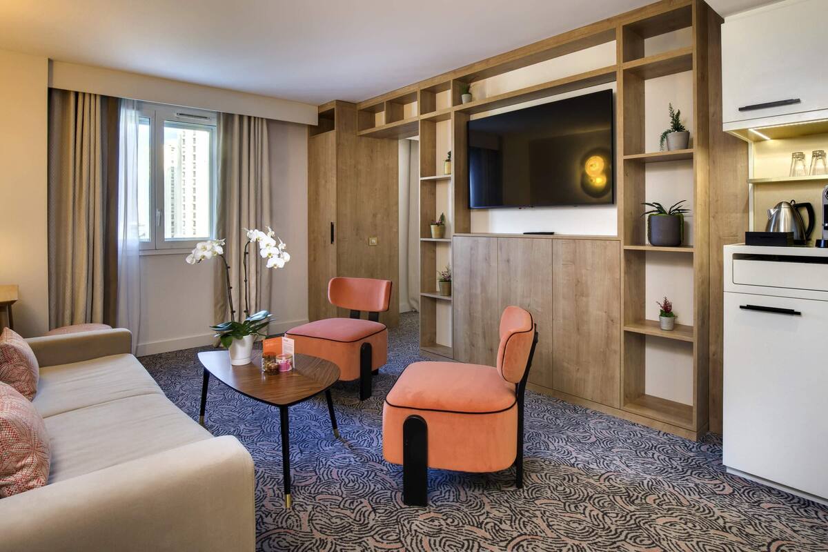 Quarto de hotel com sofá, duas cadeiras, uma mesinha, televisão, estantes de madeira e um tapete estampado.