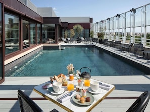 Café da manhã à beira da piscina, com bandeja de comida e bebidas, com vista para uma piscina e um edifício moderno.