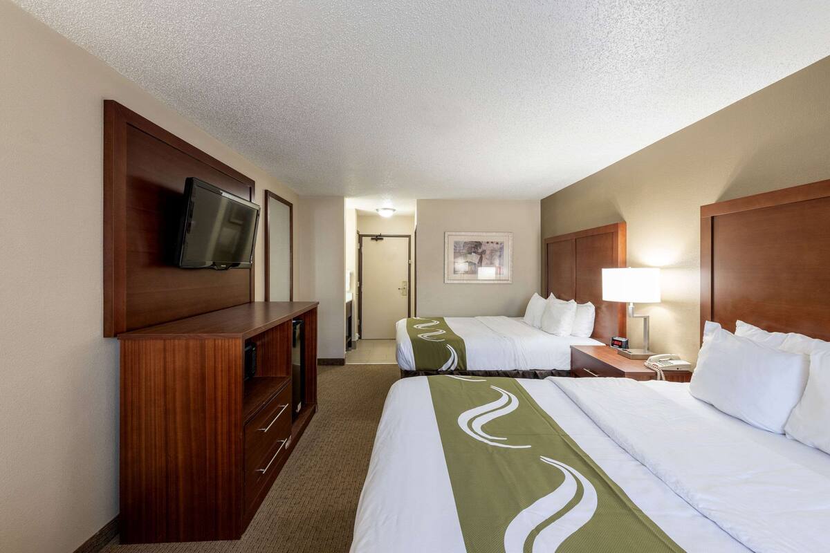 A hotel room with two beds, a TV on a wooden cabinet, a desk with a lamp, and a framed picture on the wall.