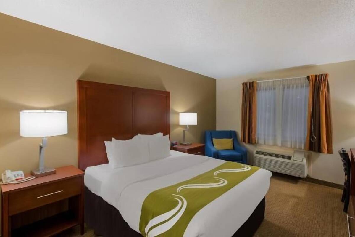 A hotel room with a bed, two bedside tables, a desk, a chair, a window with curtains, and an air conditioning unit.