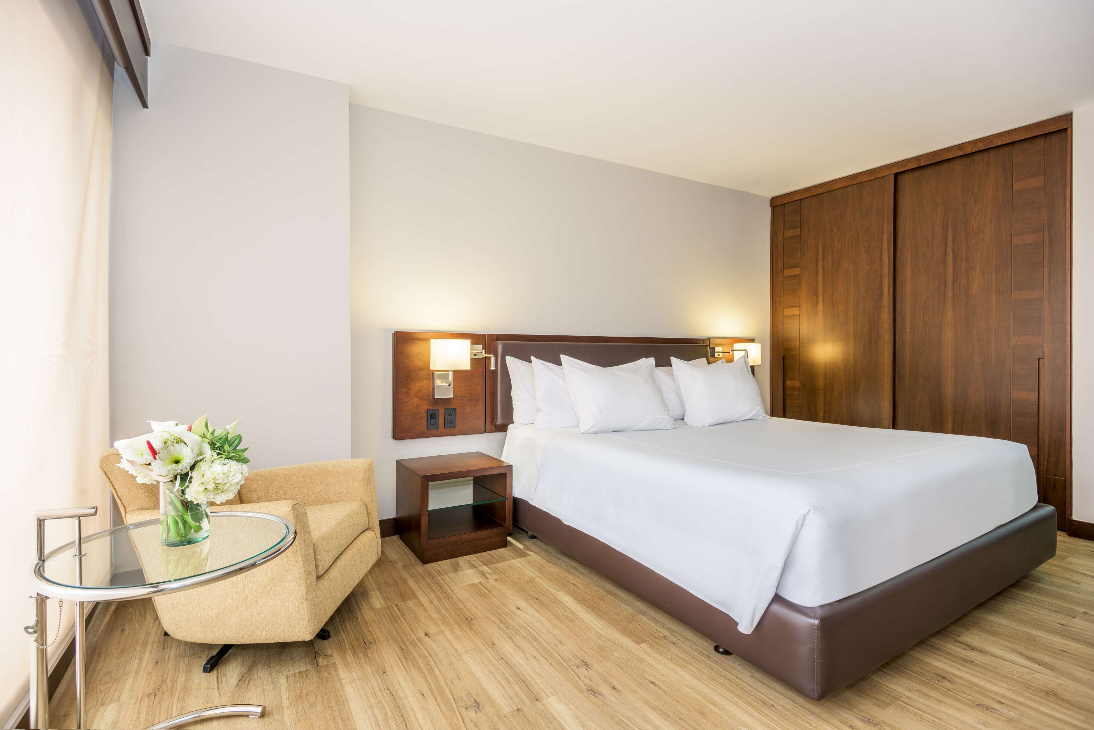 Una habitación de hotel con cama, mesita de noche, sillón y una mesa de cristal con un jarrón de flores.
