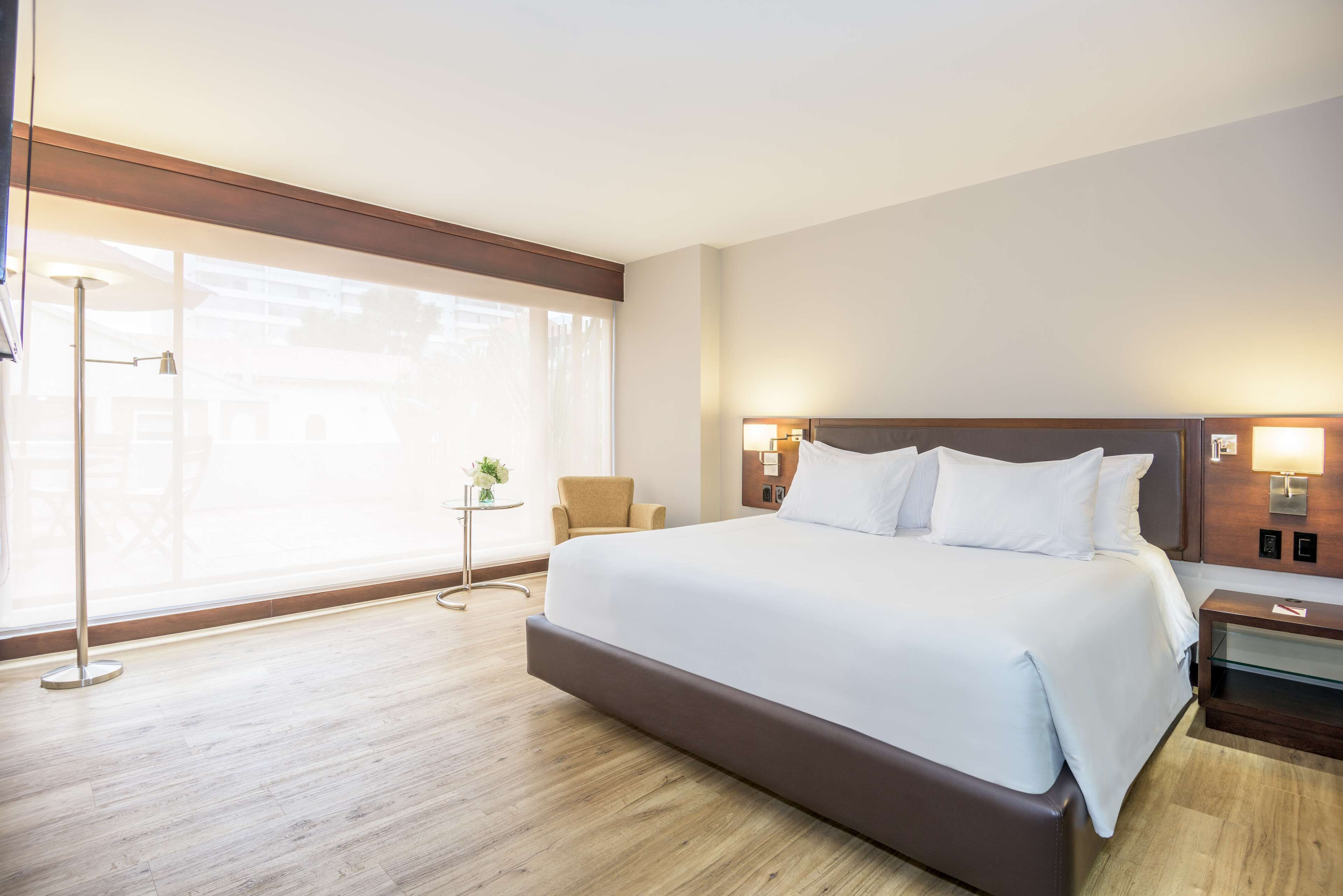 Una cama grande con sábanas blancas, una mesita de noche con lámpara y una silla cerca de una ventana con cortinas translúcidas.