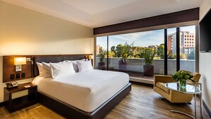 A hotel room with a large bed, a bedside table with a phone, a small round table with a vase, and a view of the city through large windows. - NH Collection Bogotá WTC Royal (Bogotá)