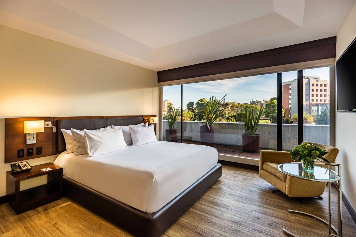 A hotel room with a large bed, a bedside table with a phone, a small round table with a vase, and a view of the city through large windows.
