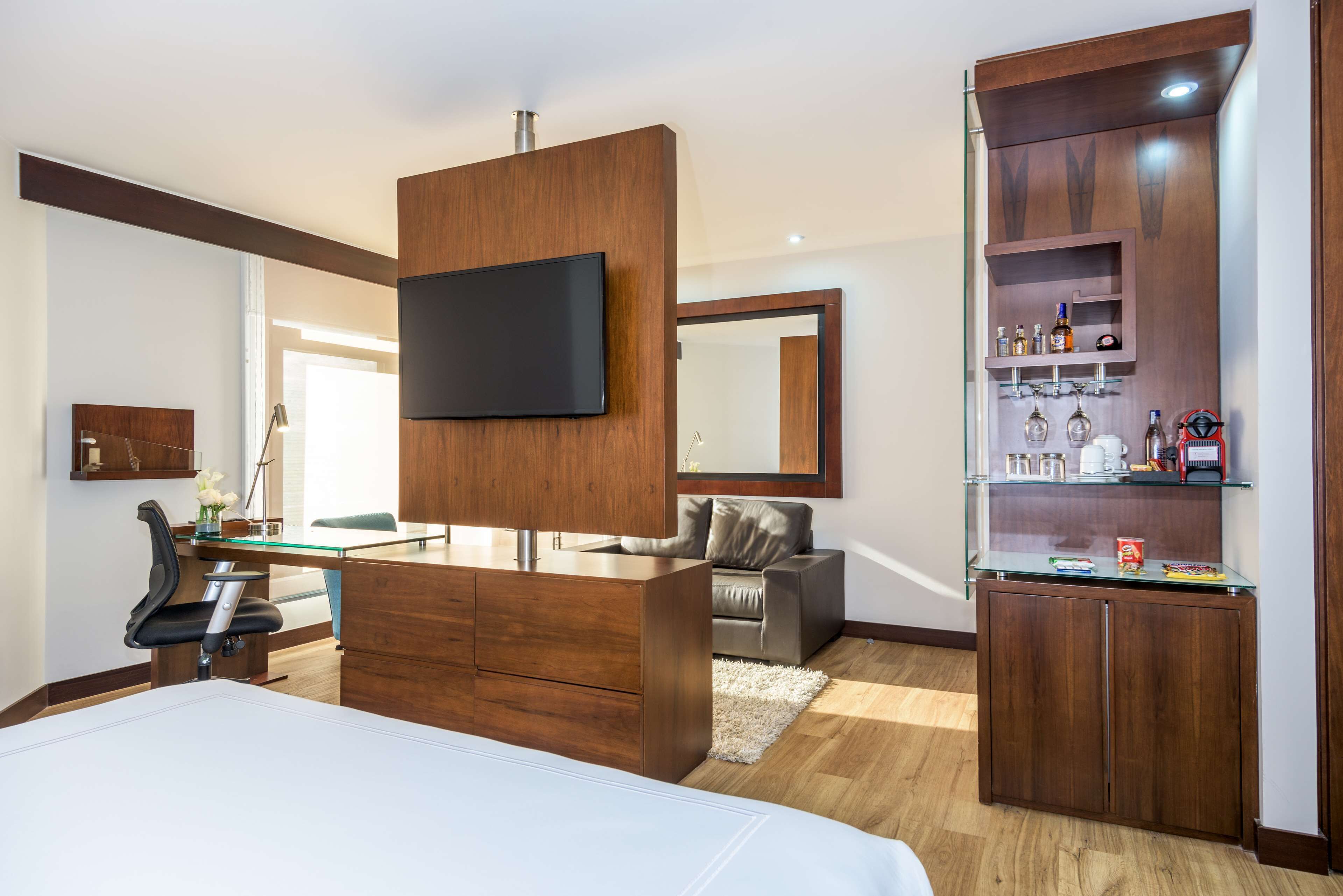 A modern bedroom with a large bed, a wooden dresser, a TV mounted on the wall, a desk, and a seating area with a chair and sofa.