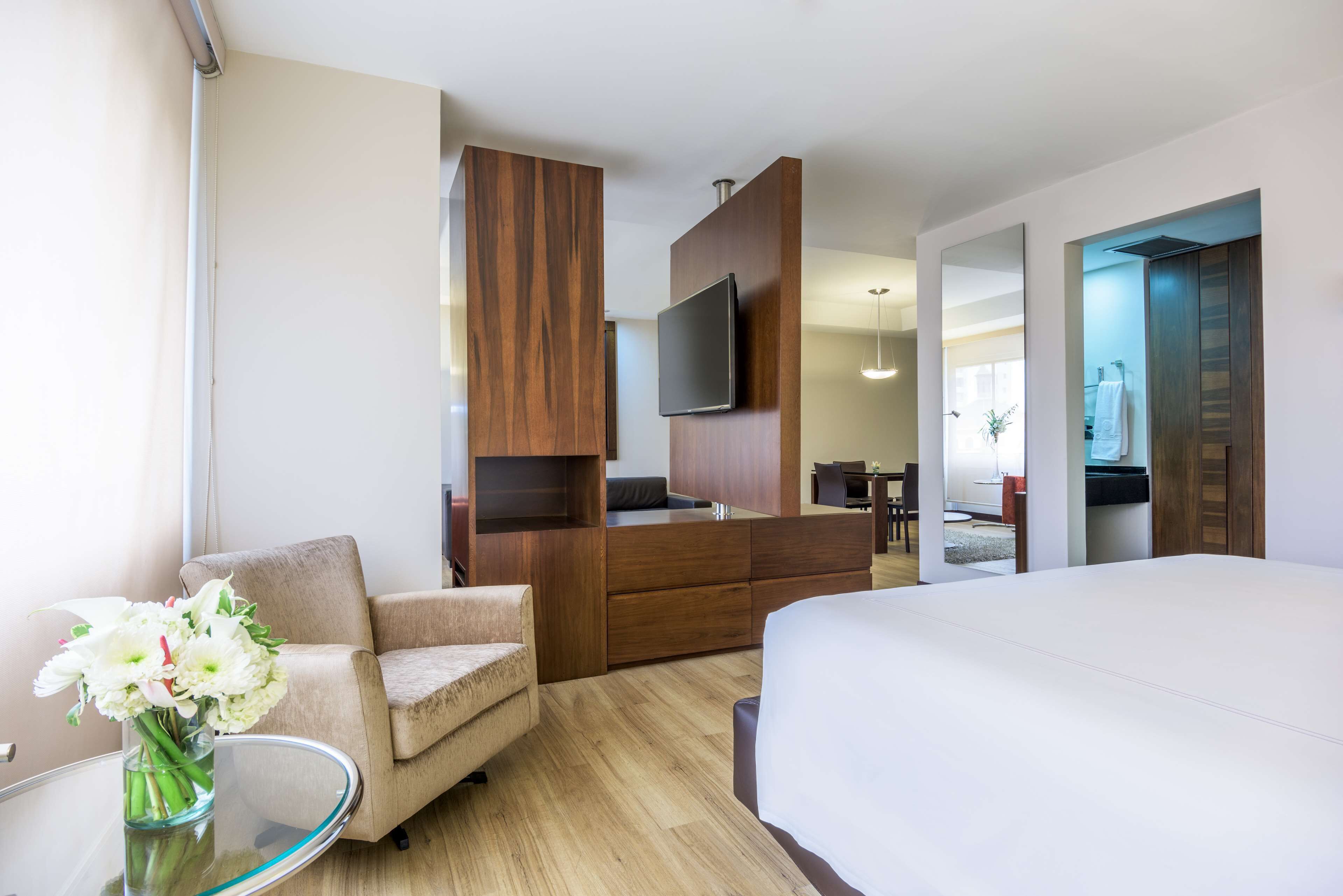 A modern hotel room with a large bed, a sofa, a TV mounted on a wooden panel, and a small table with a vase of flowers.