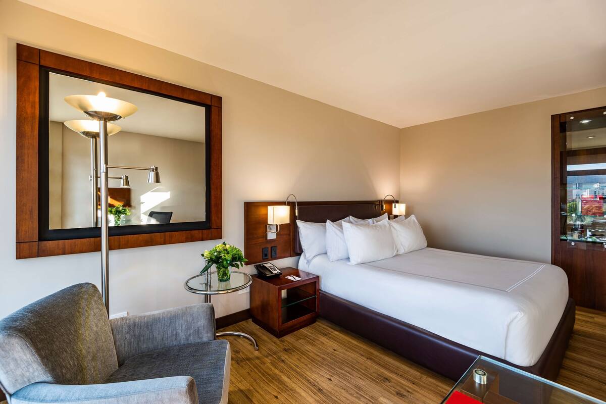 A hotel room with a large bed, a chair, a bedside table with a lamp, and a mirror on the wall.