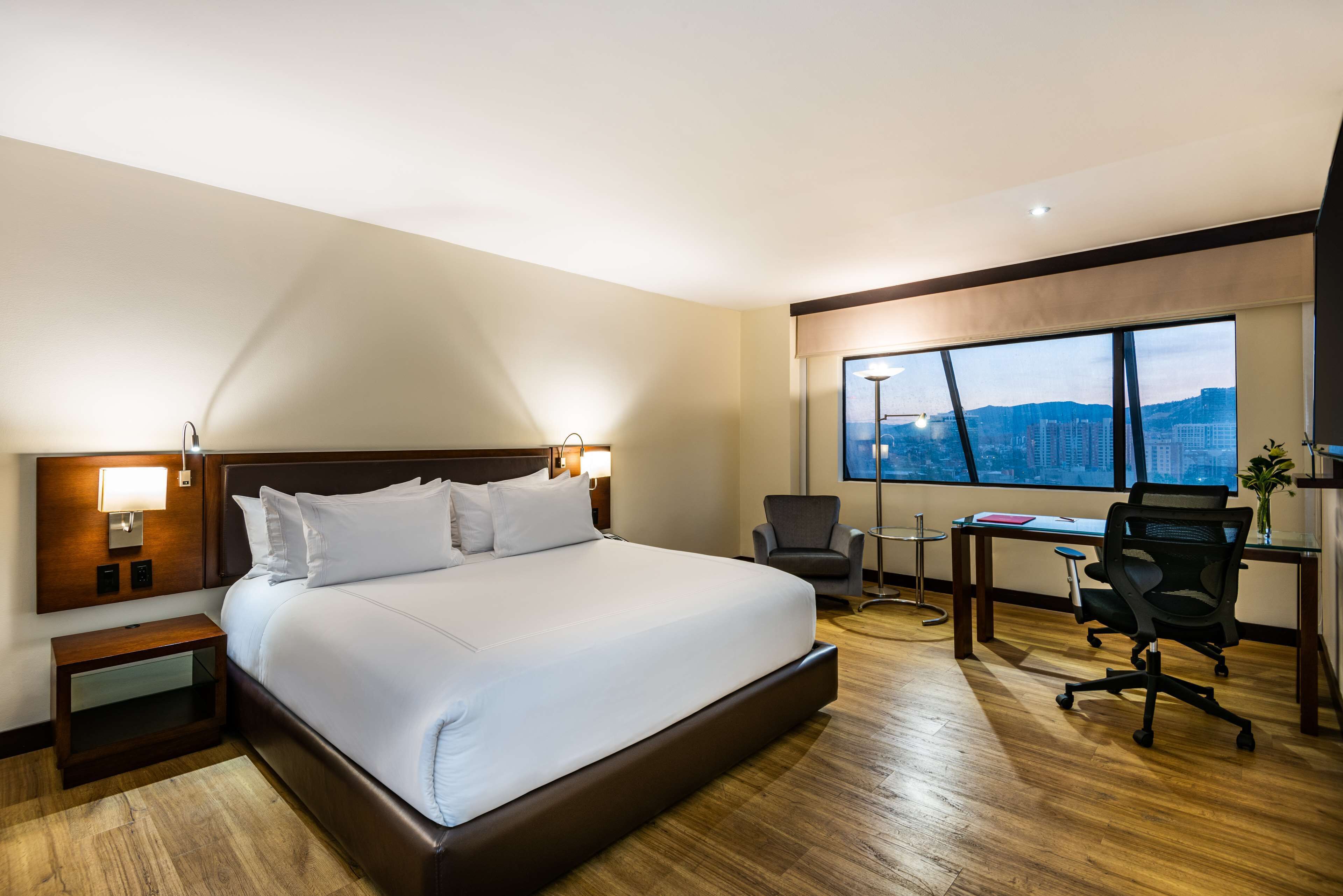 A hotel room with a large bed, a desk, a chair, and a view of a cityscape through a large window.