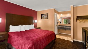 A hotel room with a large bed, a wooden headboard, a red bedspread, a nightstand, a dresser, a mirror, a sink, and a door leading to a bathroom. - Econo Lodge Cornersville I-65 (Cornersville)