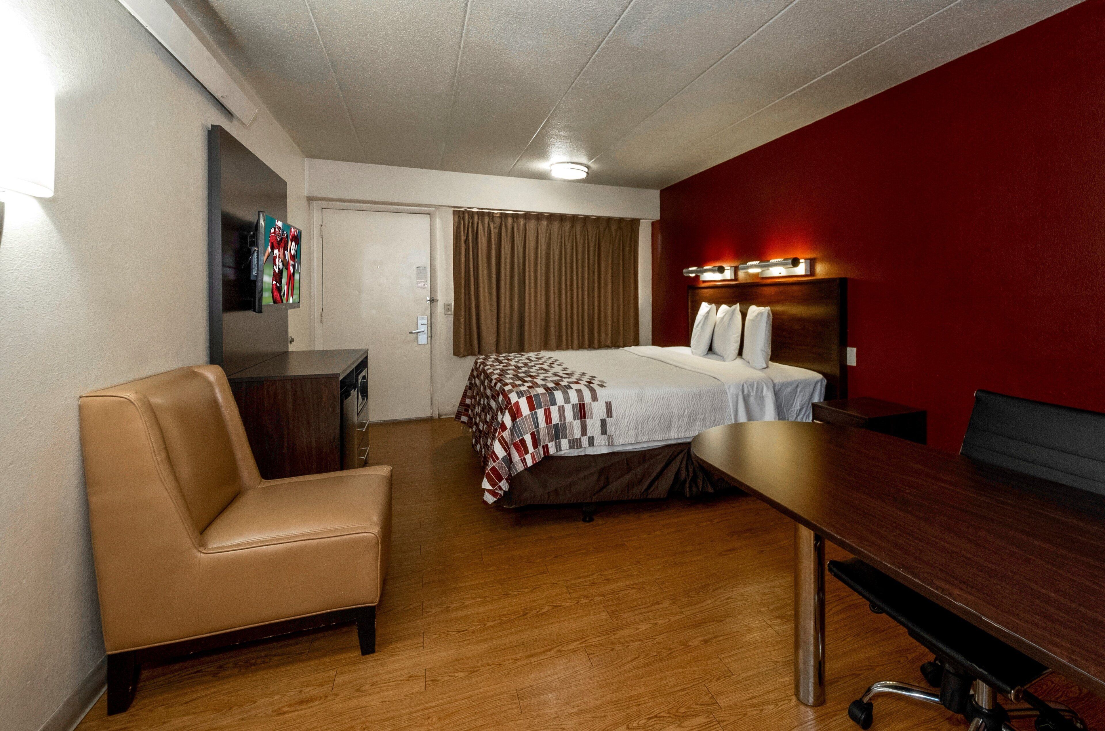 Quarto de hotel com cama, escrivaninha, cadeira, televisão e parede vermelha.