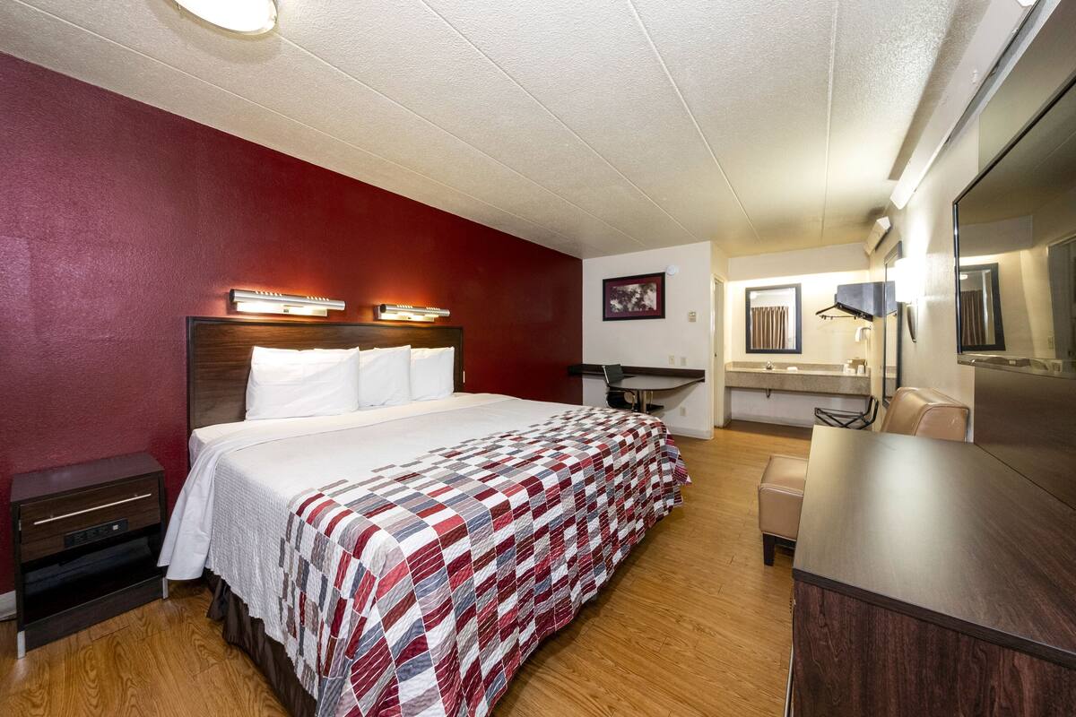 A hotel room with a large bed, a wooden nightstand, a desk, a chair, a mirror, and a bathroom with a sink and mirror.