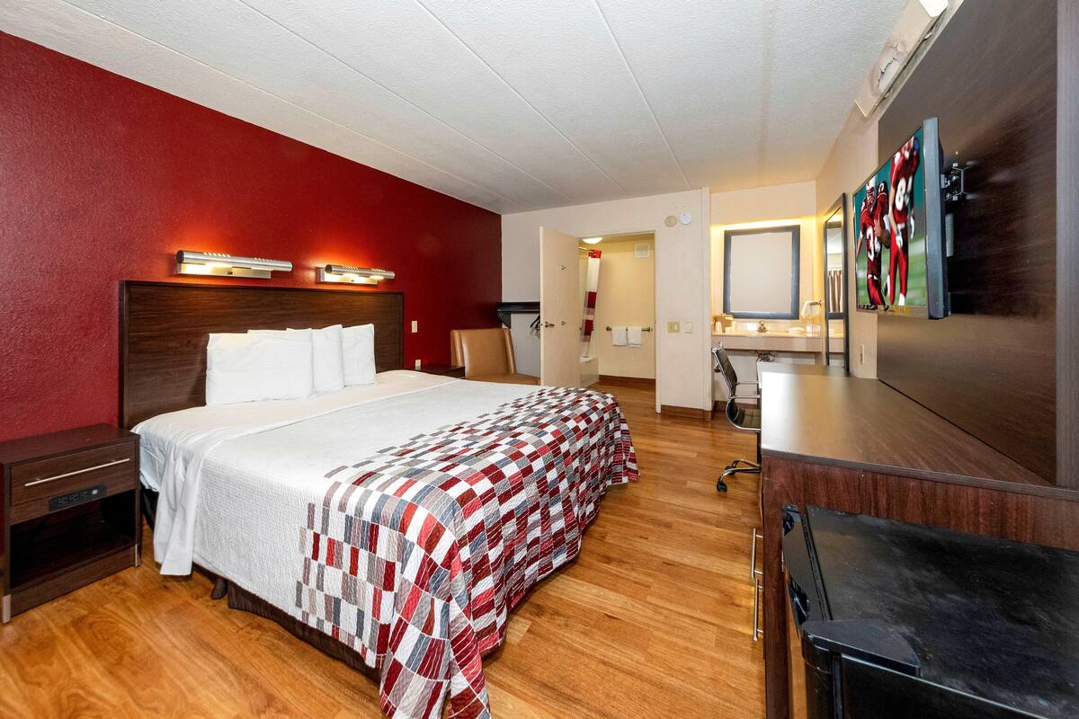 A hotel room with a large bed, a wooden nightstand, a desk with a chair, a mirror, and a TV.