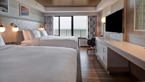 A hotel room with two beds, a TV, a desk, and a view of the sea.