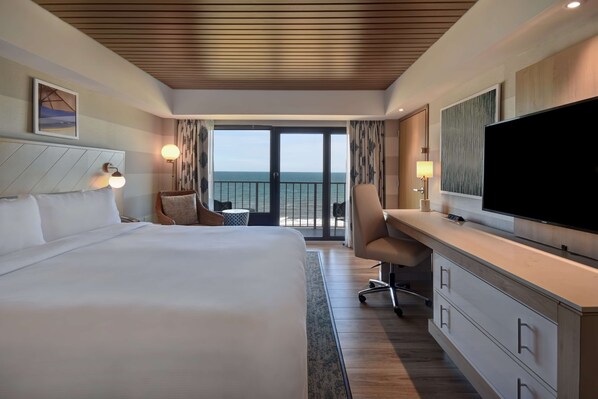 A hotel room with a large bed, a desk with a TV, a chair, and a view of the sea through a balcony.