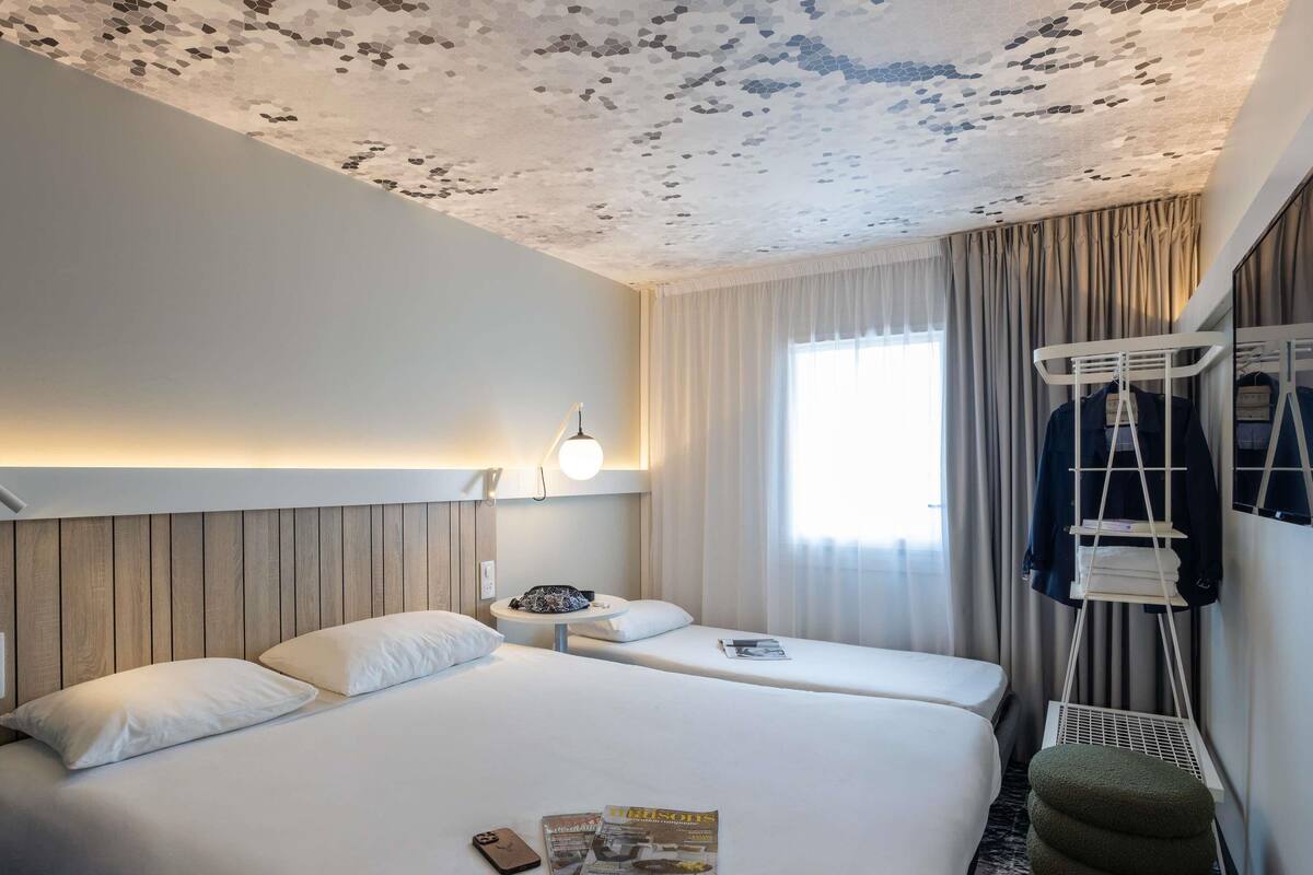 A hotel room with two beds, a round table, a lamp, a window with curtains, and a coat rack.