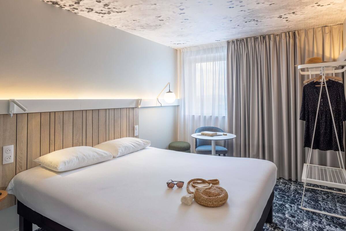 A hotel room with a bed, a small table, a chair, a hanging clothes rack, and a window with curtains.
