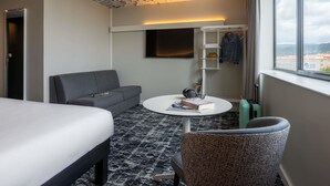 Ein Hotelzimmer mit Bett, Sofa, Couchtisch mit Büchern, Fernseher, Kleiderschrank und Blick auf die Stadt.