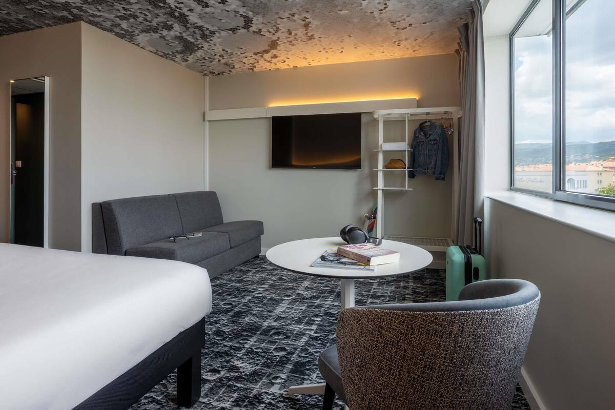 A hotel room with a bed, a sofa, a coffee table with books, a TV, a wardrobe, and a view of the city.