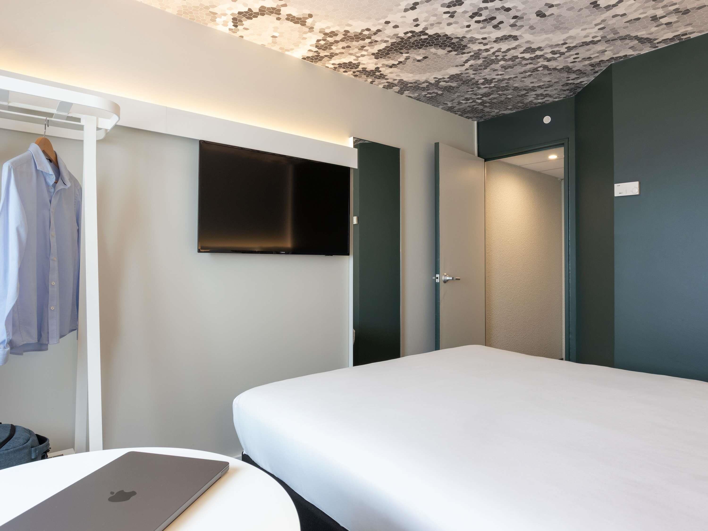 Quarto de hotel moderno com cama, televisão de tela plana, cabideiro e um laptop sobre uma mesa.