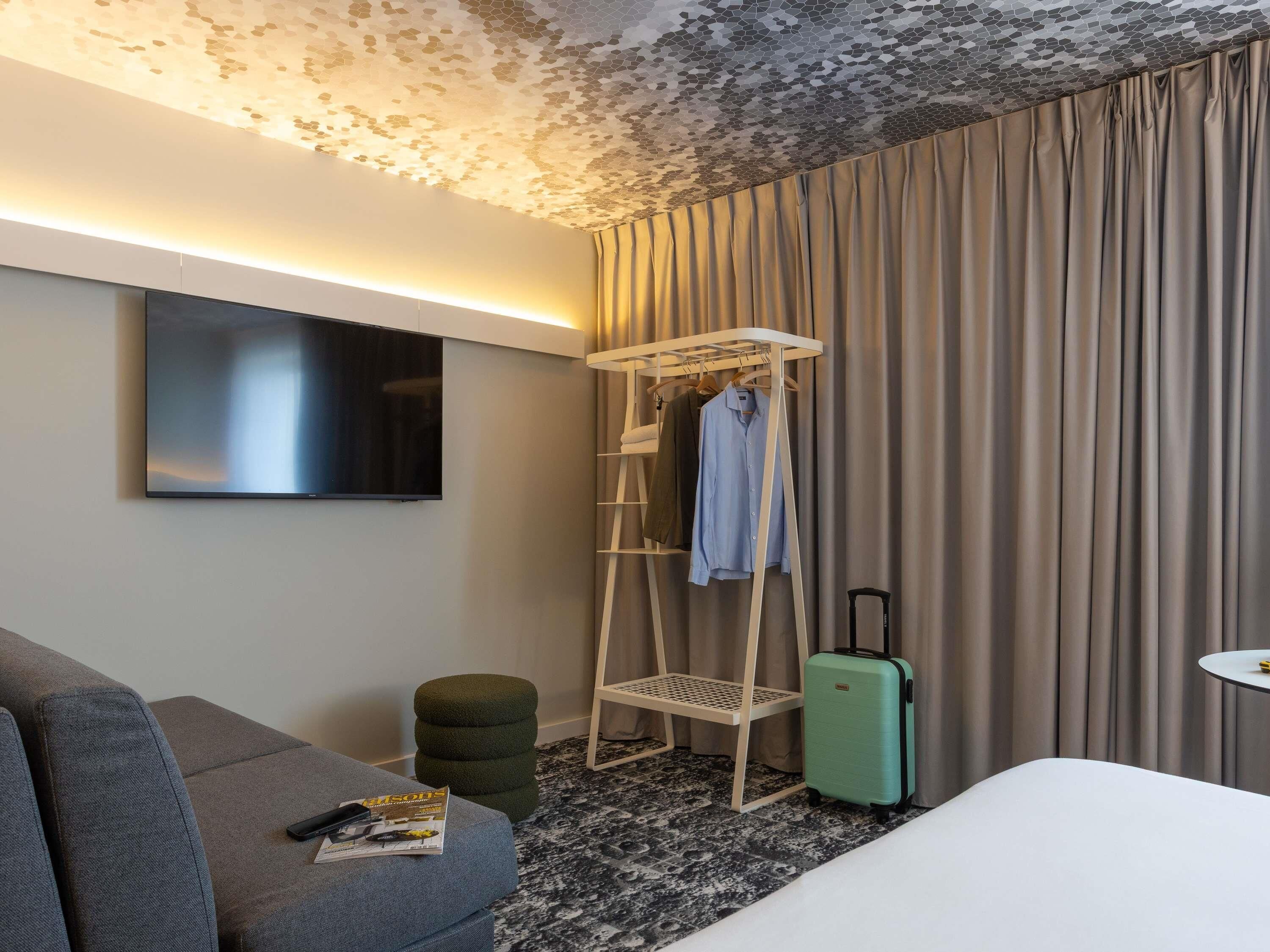 Une chambre d’hôtel moderne dotée d’une télévision à écran plat, d’un coin salon avec un canapé et un pouf, d’une armoire avec des vêtements suspendus, d’une valise et d’une petite table.