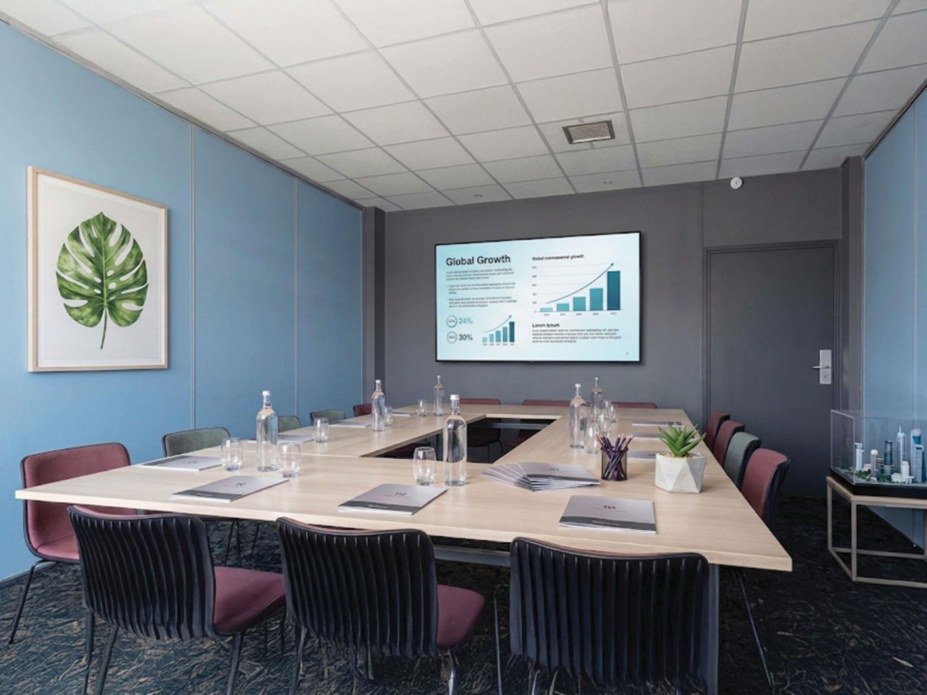 A conference room with a long table, chairs, a projector screen displaying a presentation, and a framed plant on the wall.