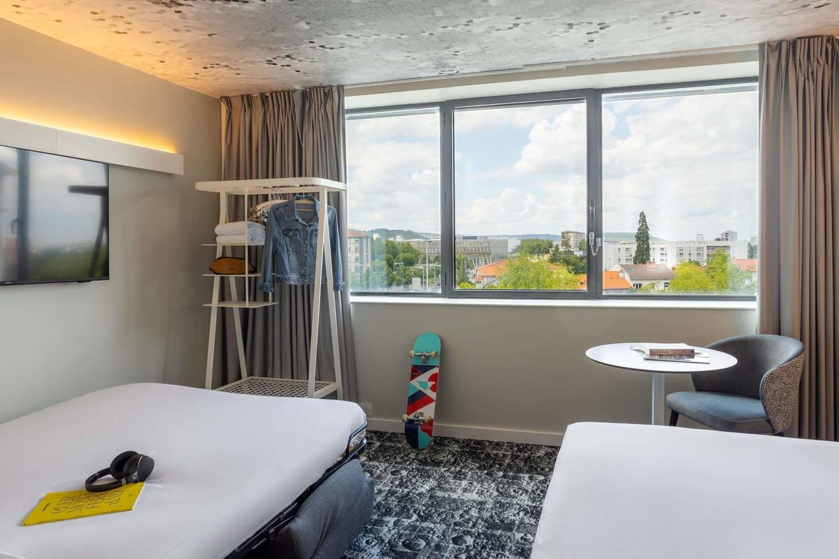 A hotel room with two beds, a TV, a small table, a chair, a shelf with clothes, and a window with a city view.