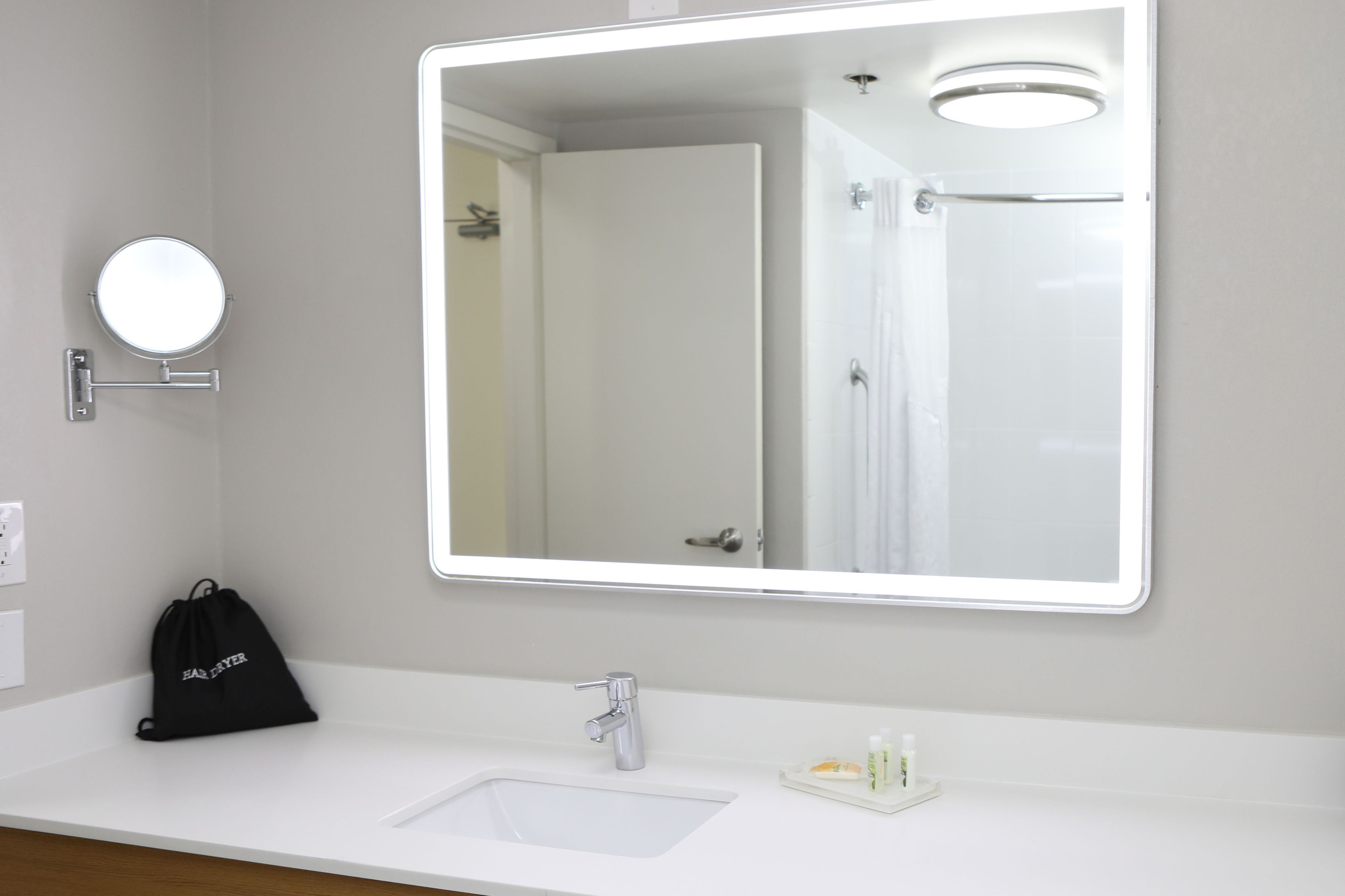 A bathroom with a large mirror, a sink, a black bag, and a small bottle on the counter.
