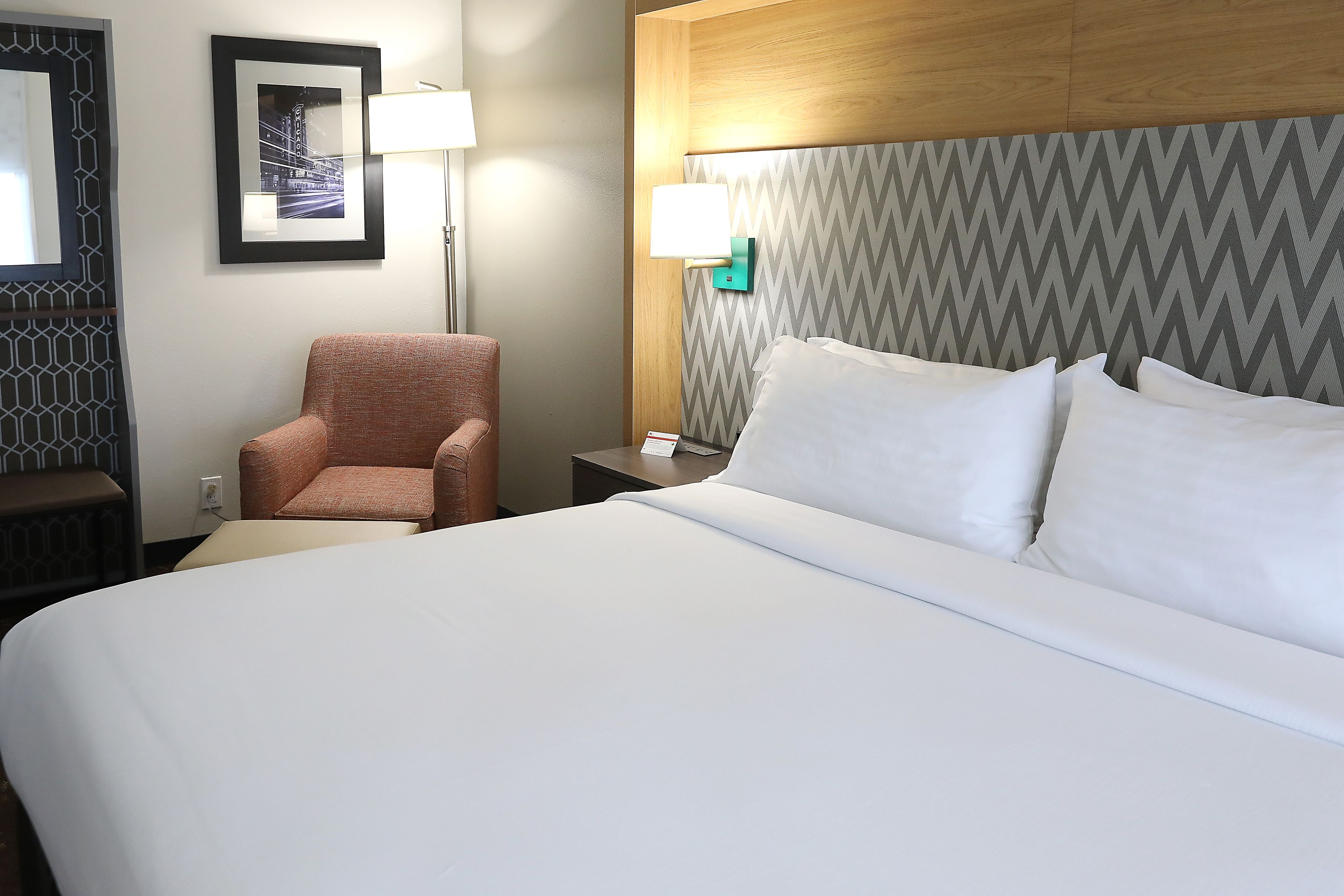 A hotel room with a large bed, a chair, a nightstand, and a wall-mounted lamp.