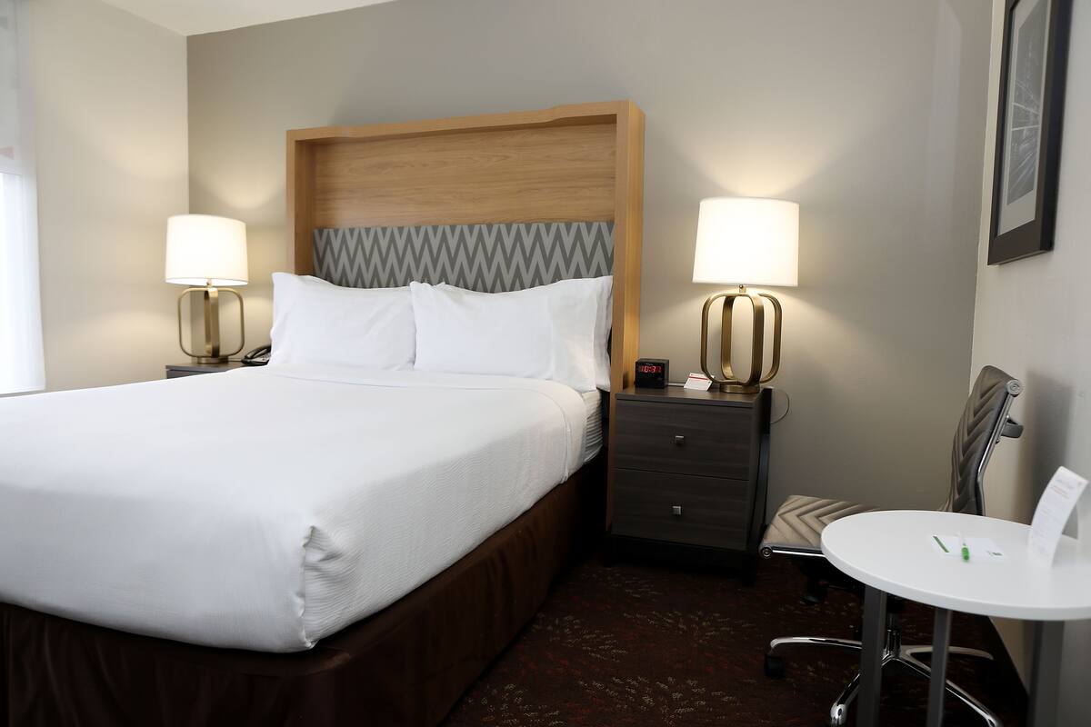 A hotel room with a bed, two bedside tables with lamps, a small desk with a chair, and a wall-mounted clock.
