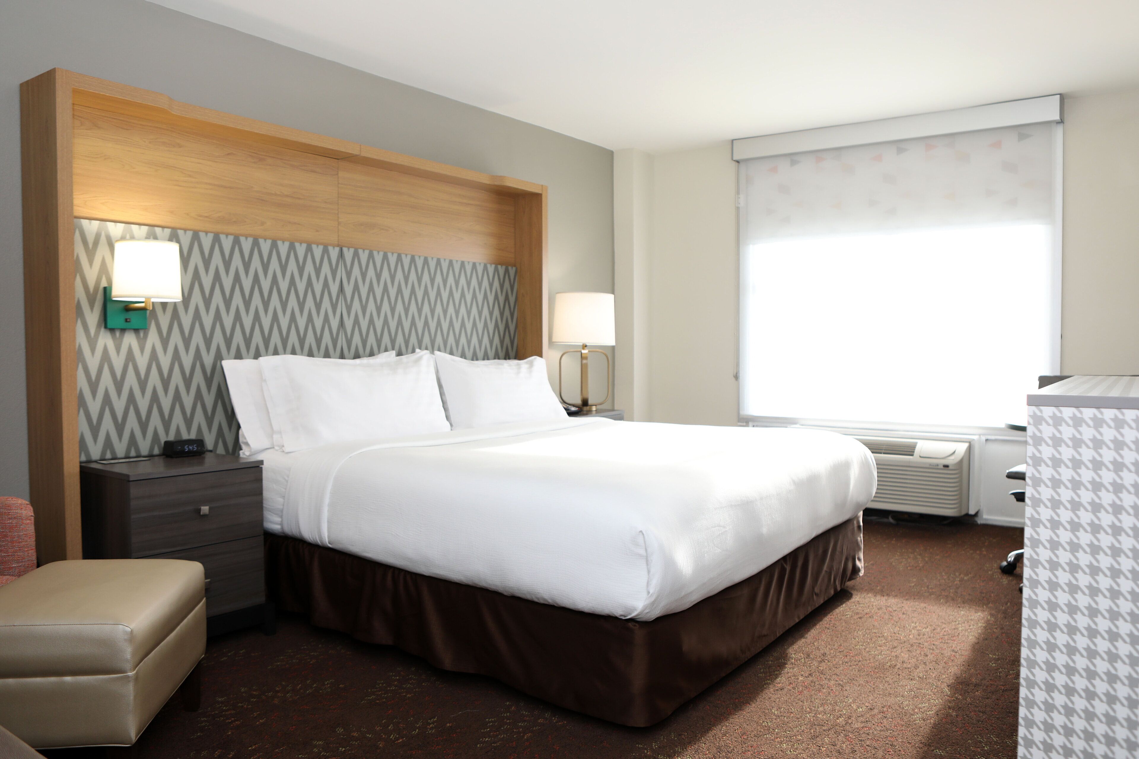 A hotel room with a large bed, a wooden headboard, two bedside tables with lamps, a bench, and a large window with a view.