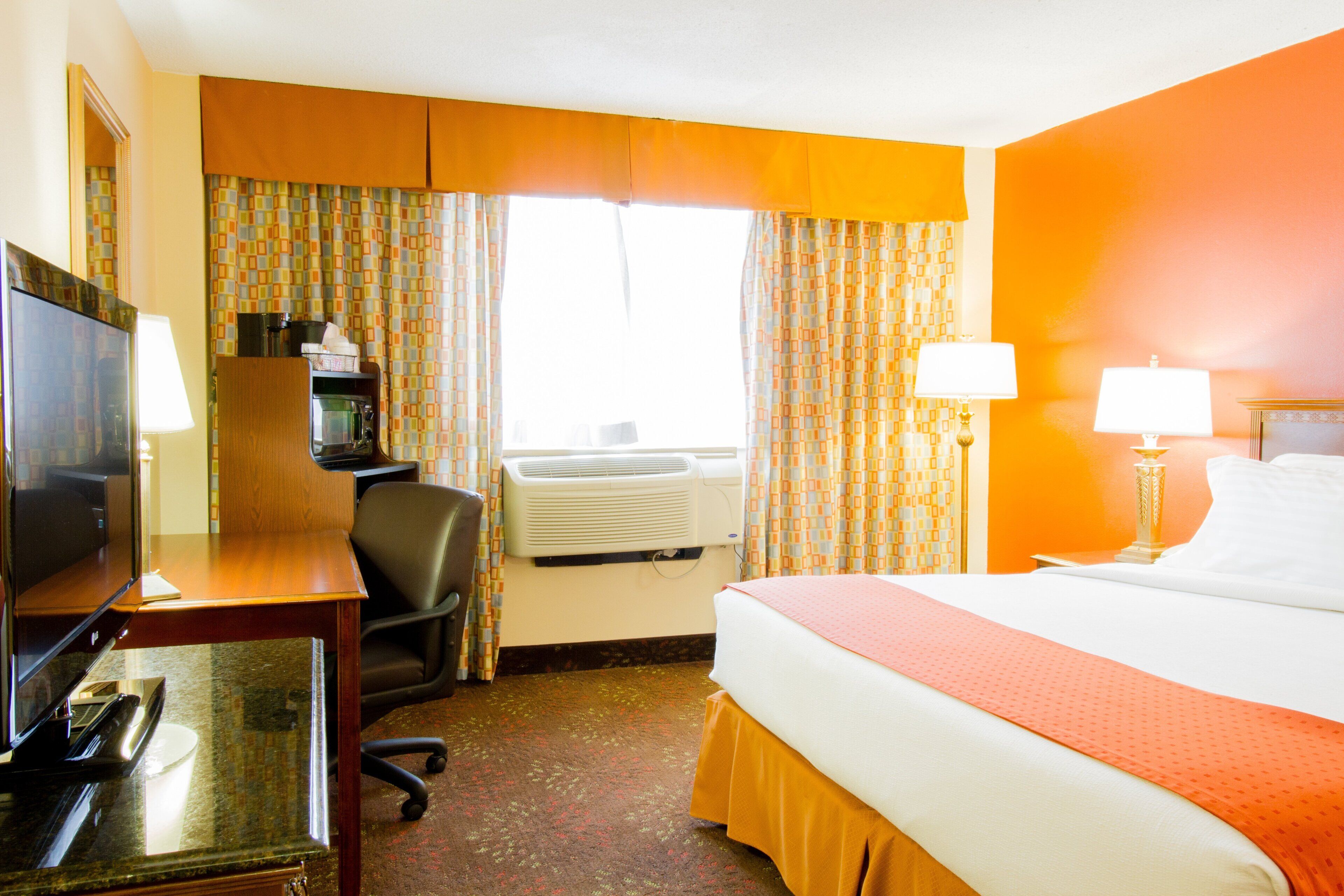 Een hotelkamer met een bed, bureau, stoel, televisie, airconditioning en gordijnen.