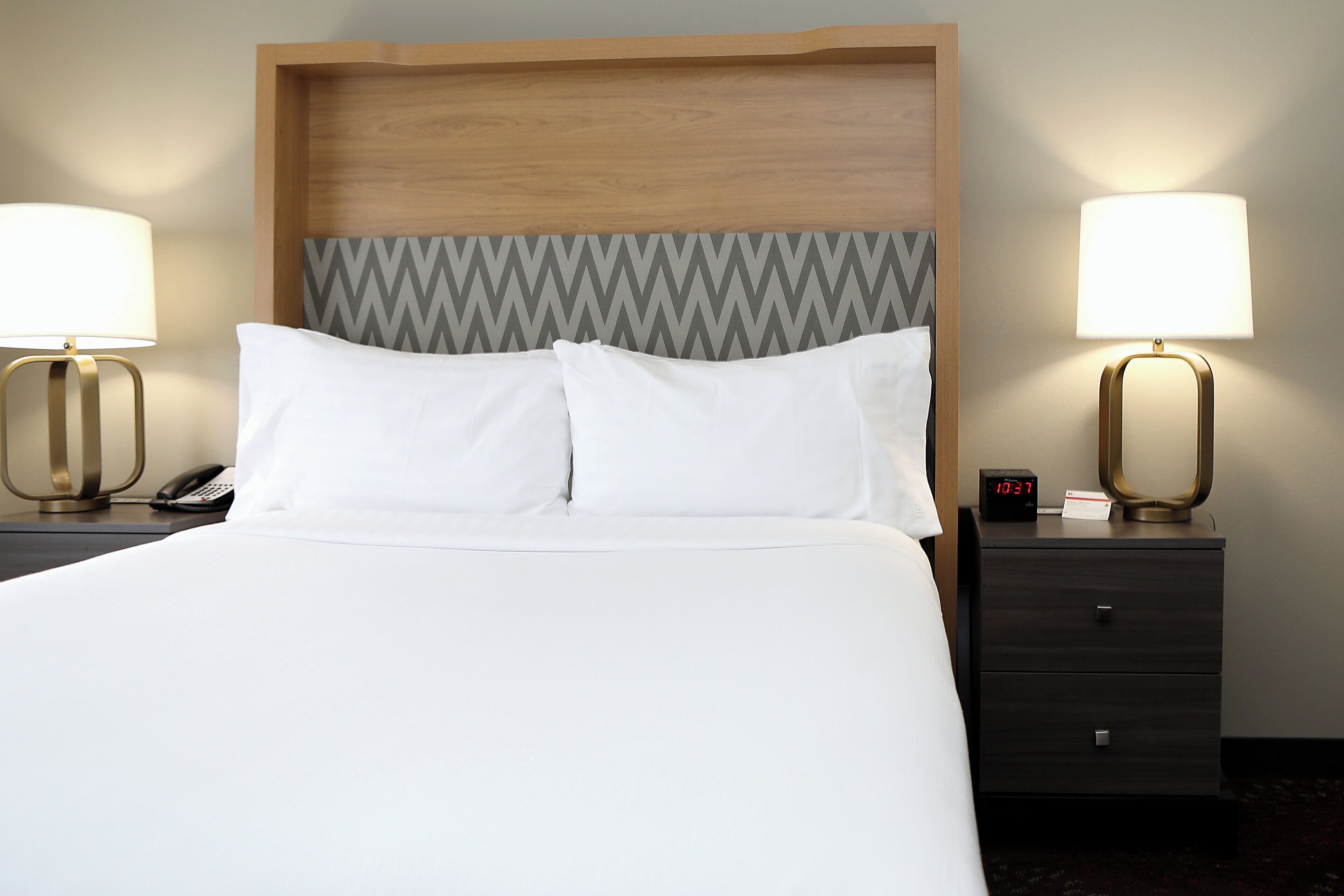 A hotel room with a bed, two bedside tables, a nightstand, and a wall-mounted clock.