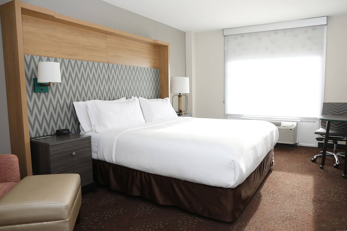 A hotel room with a bed, nightstand, lamp, and a desk with a chair.
