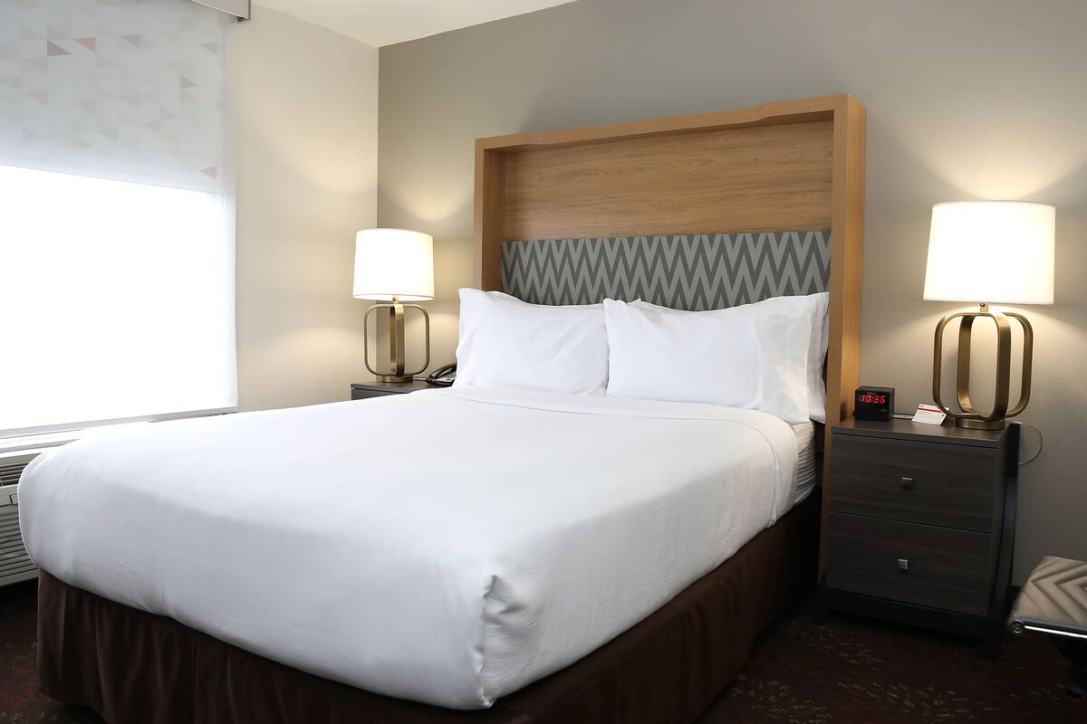 A neatly made bed with white linens, a wooden headboard with a chevron pattern, two bedside lamps, and a digital clock on the nightstand.