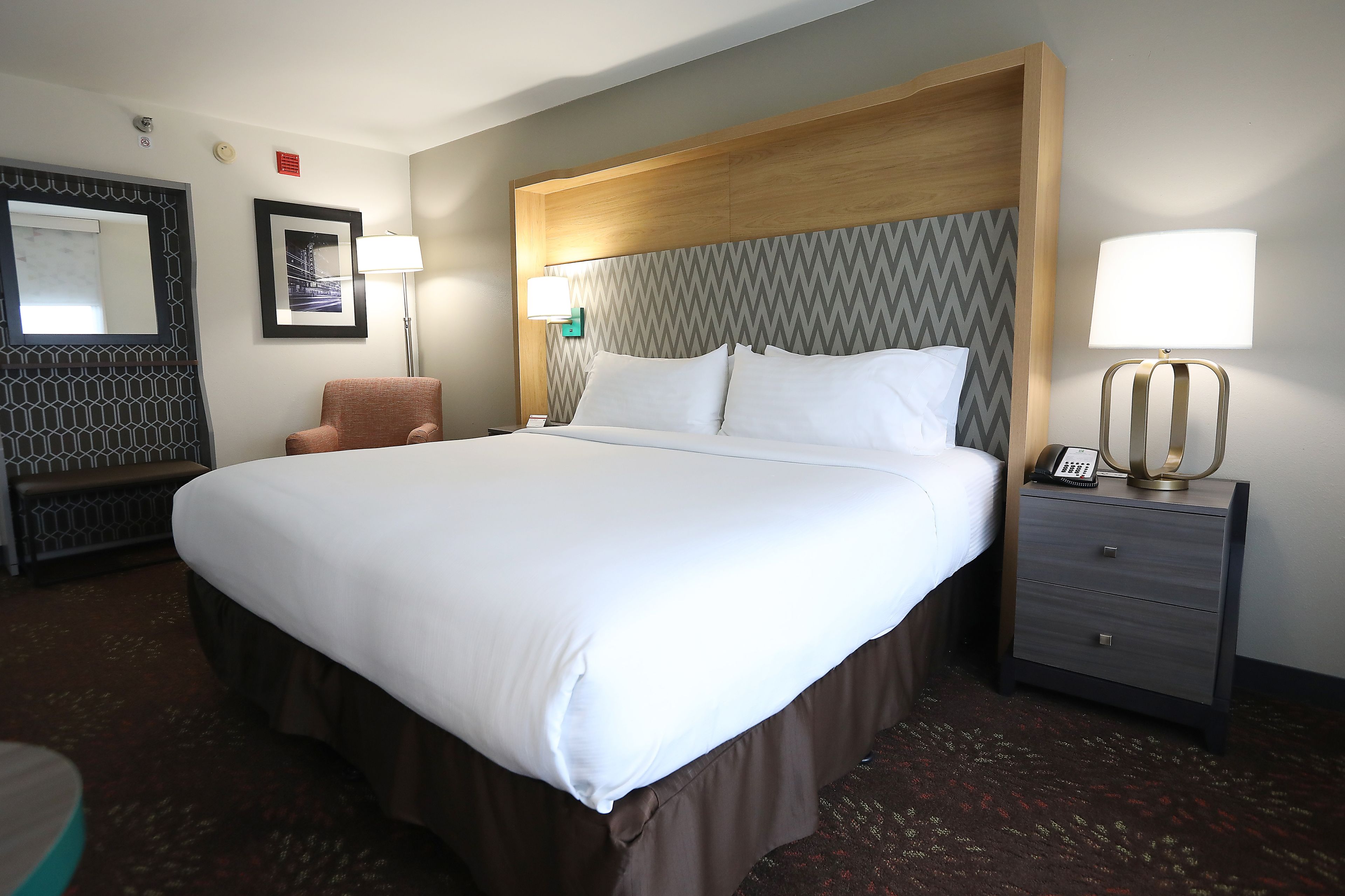 A hotel room with a large bed, a chair, a bedside table with a lamp, and a wall-mounted mirror.